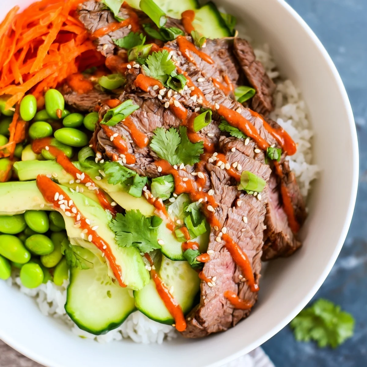 Vibrant flank steak bowl with sliced beef, fresh avocado, edamame, and drizzled spicy mayo sauce over fluffy rice