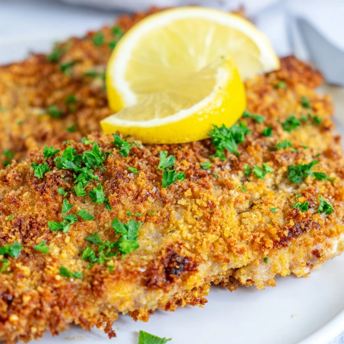 Golden crispy beef escalope Milanese garnished with fresh parsley and lemon wedges on a white plate